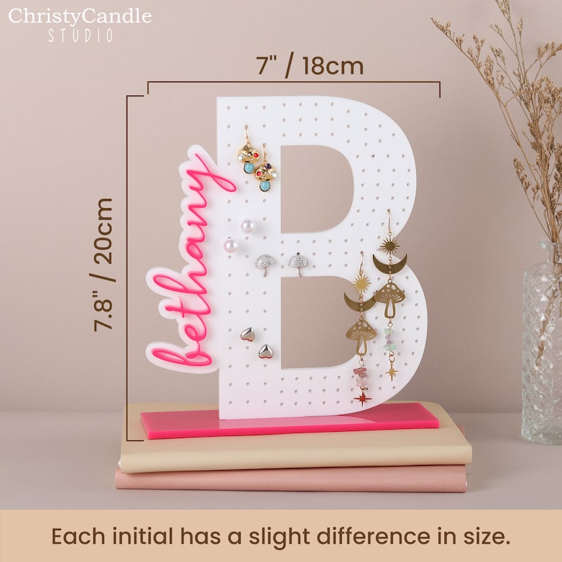 May include: White letter B earring holder with pink base. The letter B is 18 cm tall and 20 cm wide. The holder has a pink base and is decorated with the name "bethany" in pink lettering.