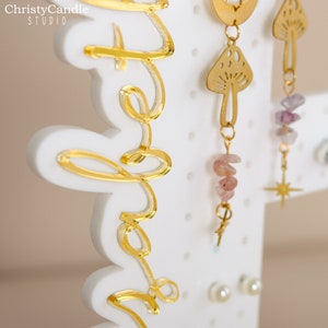 May include: White earring display stand with gold lettering that says "Hello" and gold earrings with pink and purple stones, a mushroom design, and a star design.
