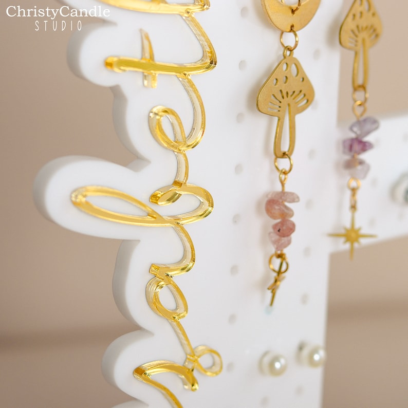 May include: White earring display stand with gold lettering that says "Hello" and gold earrings with pink and purple stones, a mushroom design, and a star design.