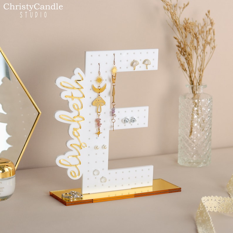 May include: White wooden earring holder shaped like the letter E with gold lettering spelling "elizabeth". The holder has a gold base and is decorated with white polka dots. The holder is displaying a variety of gold and silver earrings.