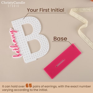 May include: White acrylic letter B with a pink base and a pink inscription that reads "bethany". The letter has a dotted surface and is designed to hold over 65 pairs of earrings.