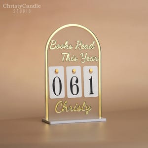 May include: A clear acrylic book counter with gold trim and a white base. The text "Books Read This Year" and "Christy" are in gold script. The counter displays the number "061".