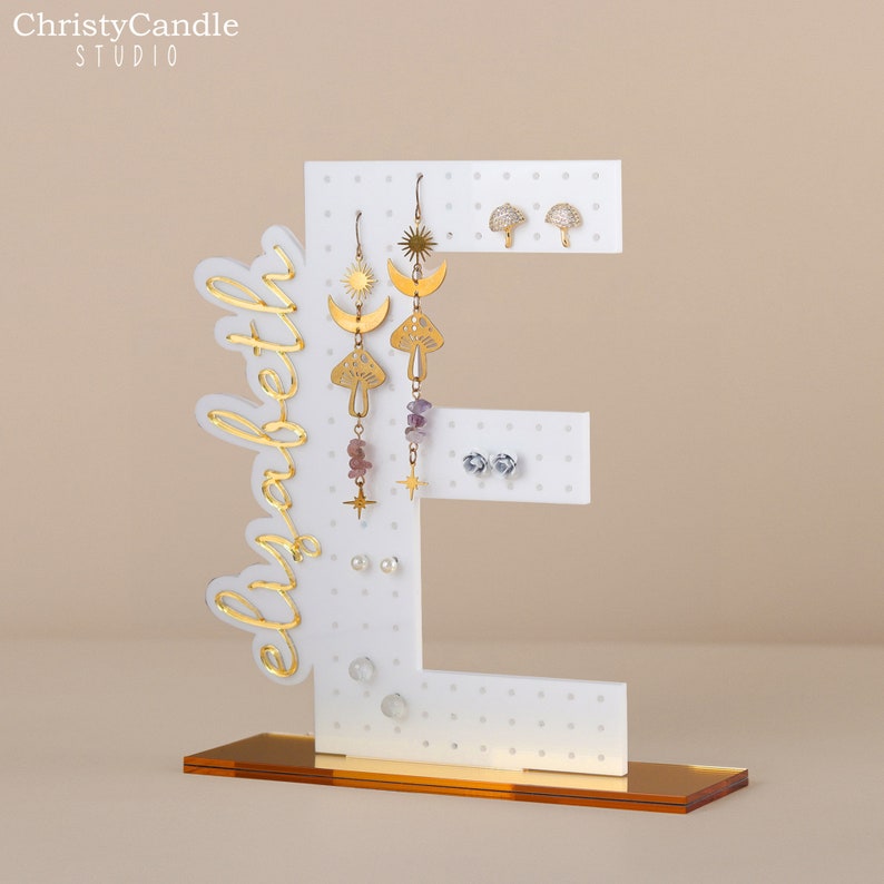 May include: White acrylic earring stand shaped like the letter E with gold lettering that says "elizabeth". The stand has multiple holes for earrings and is displayed on a gold base.