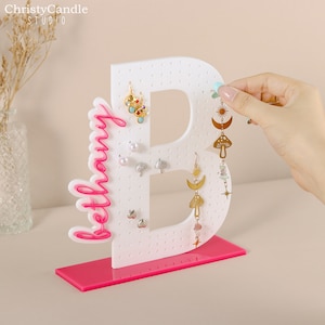 May include: White acrylic earring holder shaped like the letter B with the word "Bethany" in pink lettering. The holder has a pink base and is decorated with various earrings.