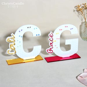 May include: Two decorative letter cutouts, a "C" and a "G", each with a name attached. The "C" has the name "Carol" in gold script, and the "G" has the name "Grace" in pink script. Both letters are white with a dotted pattern and sit on colored bases.