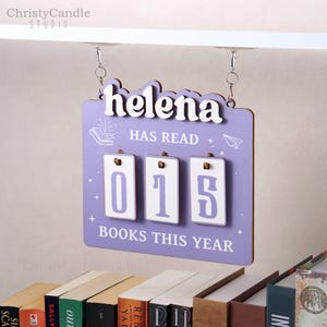 May include: A lavender wooden sign with the name "Helena" in white script. The sign displays "HAS READ" and a counter showing "015 BOOKS THIS YEAR". It hangs from silver chains and is designed for tracking reading progress.