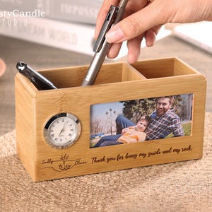 Könnte beinhalten: Ein hölzerner Schreibtisch-Organizer mit einem Bilderrahmen, einer kleinen Uhr und zwei Fächern. Der Bilderrahmen zeigt ein Foto von einem Vater und seinem Sohn. Der Organizer ist mit dem Text "Daddy Oliver Thank you for being my guide and my rock." graviert.
