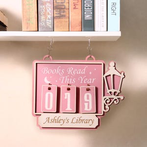 Puede incluir: Letrero de madera rosa con el texto "Books Read This Year" y los números "019". El letrero también incluye el texto "Ashley's Library" y una linterna decorativa. El letrero está colgado de una estantería con libros.
