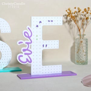 May include: A white earring holder in the shape of the letter "E" with a purple base and the name "Evie" in purple script. The holder has small holes for earrings and is decorated with purple bow and green gem accents. The Christy Candle Studio logo is in the top left corner.