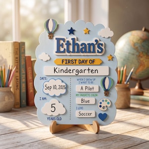 Könnte beinhalten: Ein hellblaues Holzschild für den ersten Kindergartentag mit dem Namen Ethan, einem Heißluftballon, Sternen und Wolken. Es enthält Felder für das Datum 10. Sep. 2025, das Alter 5, den Berufswunsch Pilot, die Lieblingsfarbe Blau und das Hobby Fußball.
