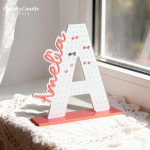 May include: A white, letter-shaped earring holder with the name "Amelia" in coral script. The holder has small holes for earrings and sits on a coral base. The Etsy shop name "ChristyCandle Studio" is in the top left corner.