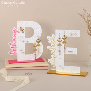 May include: White wooden letter B and E earring holders with pink and gold bases. The letters are decorated with the words "Bethany" and "Elizabeth" in gold script. The earring holders have multiple holes for displaying earrings.
