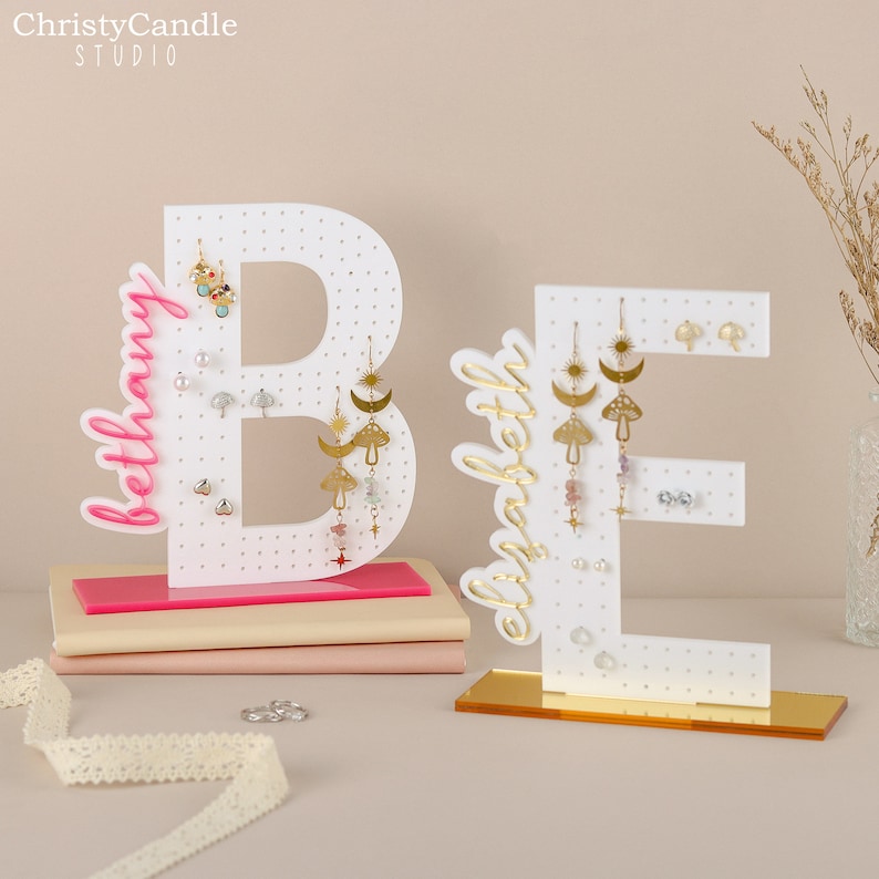 May include: White wooden letter B and E earring holders with pink and gold bases. The letters are decorated with the words "Bethany" and "Elizabeth" in gold script. The earring holders have multiple holes for displaying earrings.