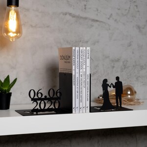 May include: Black metal bookends shaped like a couple holding hands. The bookends are holding a stack of books with titles in various languages. The date 06.06.2020 is displayed on the bookend in the foreground.