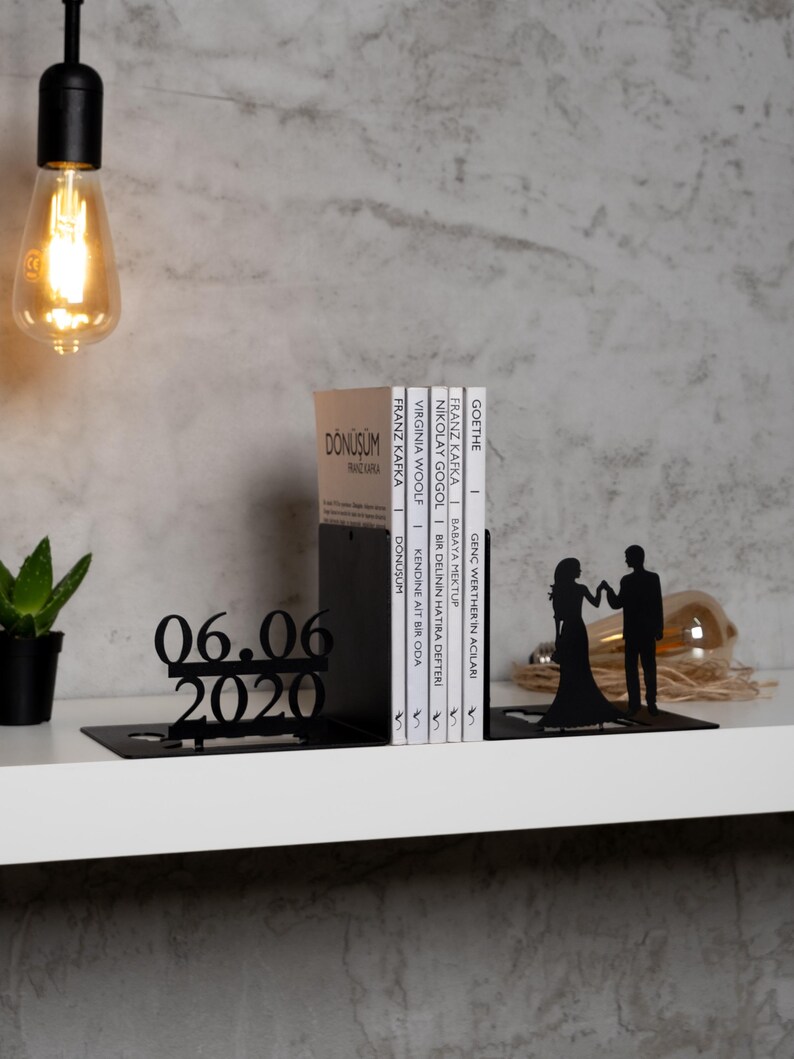 May include: Black metal bookends shaped like a couple holding hands. The bookends are holding a stack of books with titles in various languages. The date 06.06.2020 is displayed on the bookend in the foreground.