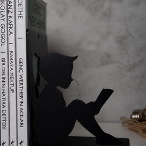 May include: Black metal bookends shaped like a silhouette of a child sitting and reading a book. The bookends are holding up three books with titles in Turkish.