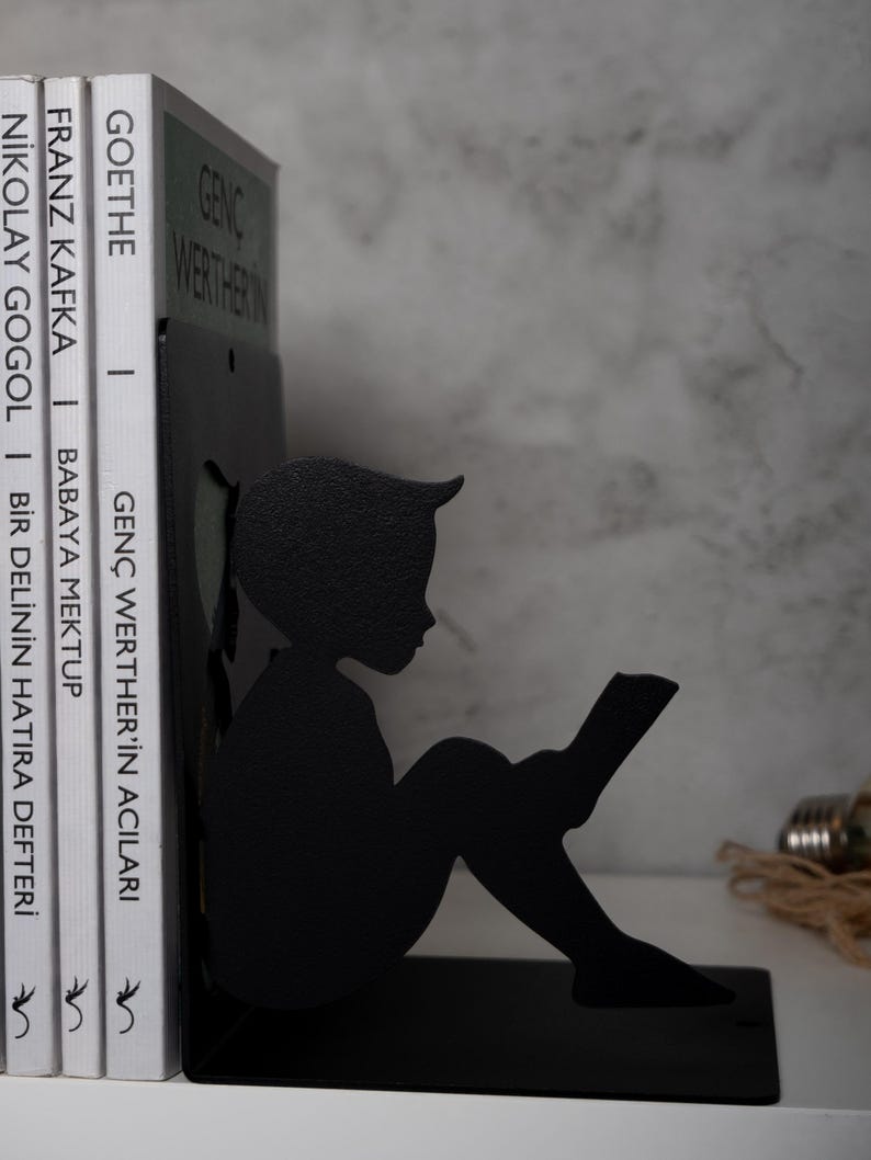 May include: Black metal bookends shaped like a silhouette of a child sitting and reading a book. The bookends are holding up three books with titles in Turkish.