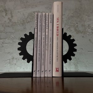 Può includere: Due ferma libri in metallo nero a forma di ingranaggi che tengono una fila di libri con copertine bianche e testo nero. I titoli dei libri includono "Yes, I Would", "I dolori del giovane Werther", "Il diario di un pazzo", "Mrs. Dalloway" e "La metamorfosi".