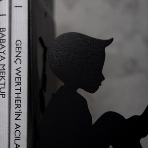 May include: Black metal bookends shaped like a silhouette of a person reading a book. The bookends are holding up two books with the titles "GEN&Ccedil; WERTHER'İN ACILARI" and "BABAYA MEKTUP".