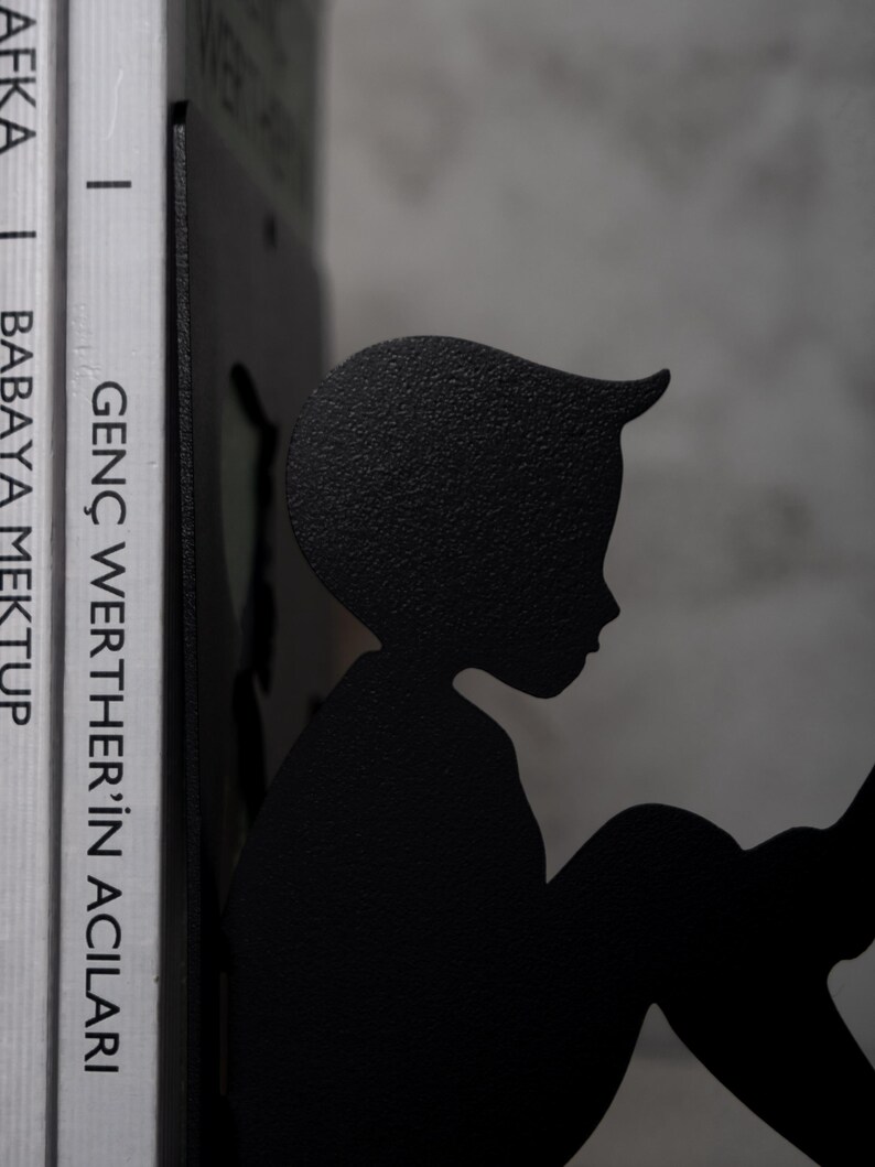 May include: Black metal bookends shaped like a silhouette of a person reading a book. The bookends are holding up two books with the titles "GEN&Ccedil; WERTHER'İN ACILARI" and "BABAYA MEKTUP".
