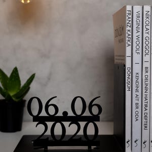 May include: Black metal bookends with a date, 06.06.2020, displayed on top. The bookends are holding up three books with titles in a foreign language.