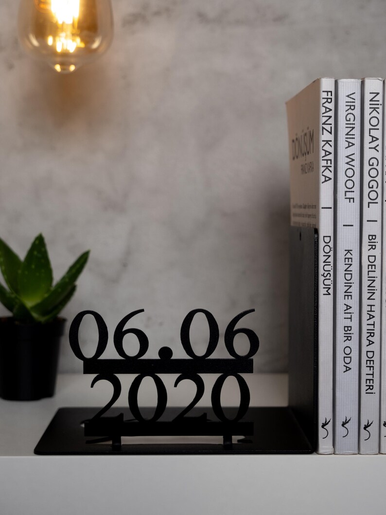May include: Black metal bookends with a date, 06.06.2020, displayed on top. The bookends are holding up three books with titles in a foreign language.