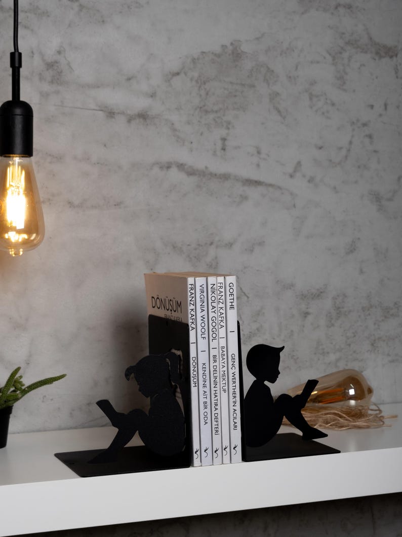 May include: Black metal bookends shaped like children reading. The bookends are holding a stack of books with titles in German and English. The titles include "Donusum", "Franz Kafka", "Virginia Woolf", "Nikolay Gogol", "Babaya Mektub", "Genc Werther'in Acıları", and "Goethe 1".