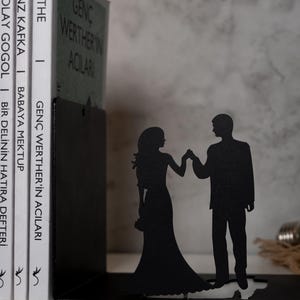 May include: Black metal bookends shaped like a couple holding hands. The bookends are holding up a stack of books with titles in German and English.