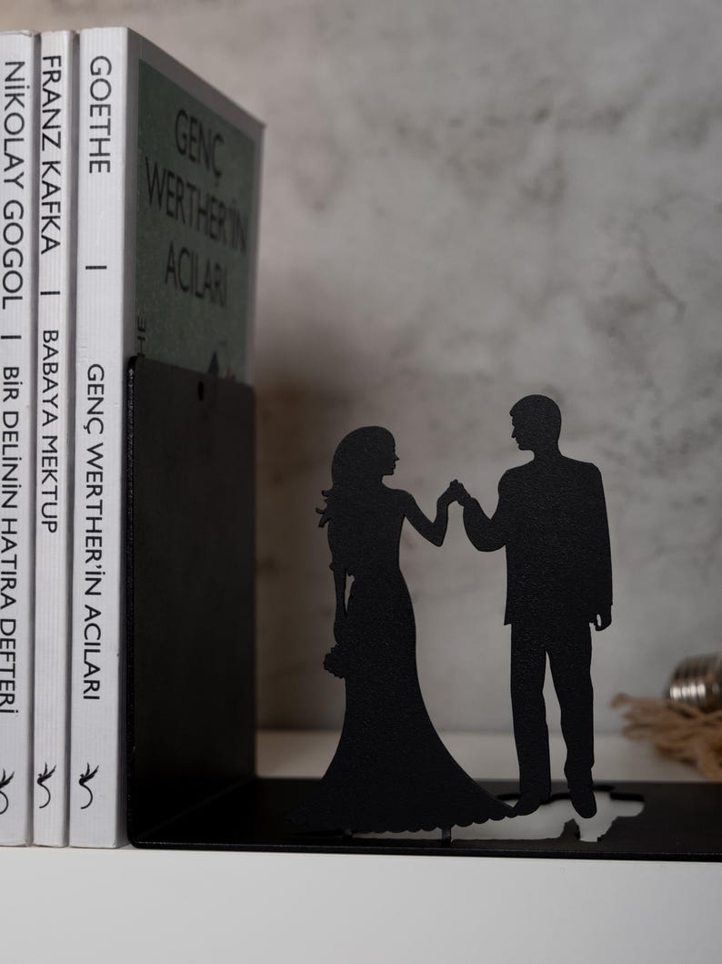 May include: Black metal bookends shaped like a couple holding hands. The bookends are holding up a stack of books with titles in German and English.