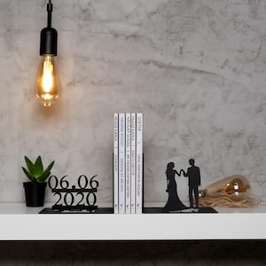 May include: Black metal bookends shaped like a couple holding hands. The bookends are holding a stack of books with white covers. The date 06.06.2020 is displayed on a black metal stand.