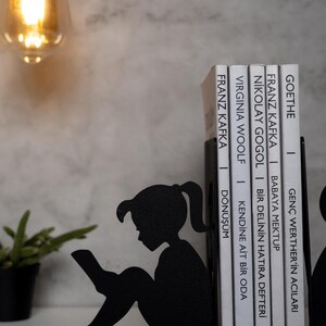 May include: Black metal bookends shaped like a girl sitting and reading a book. The bookends are holding up a stack of books with white covers and black text. The text on the books includes the titles "Goethe", "Franz Kafka", "Nikolai Gogol", "Virginia Woolf", and "Franz Kafka".