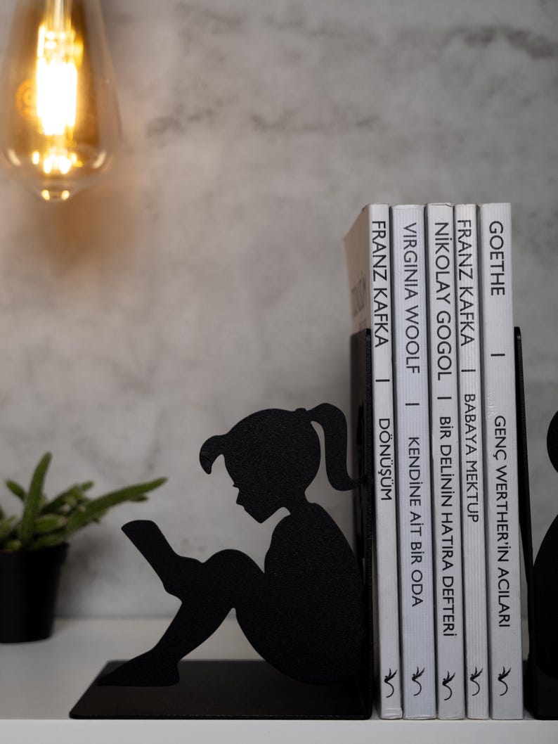 May include: Black metal bookends shaped like a girl sitting and reading a book. The bookends are holding up a stack of books with white covers and black text. The text on the books includes the titles "Goethe", "Franz Kafka", "Nikolai Gogol", "Virginia Woolf", and "Franz Kafka".