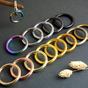 Puede incluir: Una colección de anillos de cierre redondos de metal en varios colores, incluyendo plata, oro, negro y un acabado iridiscente arcoíris. Los anillos están dispuestos sobre una superficie oscura, mostrando sus diferentes tonos y acabados. Algunos anillos están unidos a un palo de madera.
