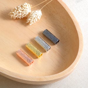 Puede incluir: Cinco cuentas de metal en forma de barril en diferentes colores: oro rosa, oro, plata y dos tonos de gris. Las cuentas tienen una superficie texturizada y están dispuestas en una fila sobre una superficie de madera.