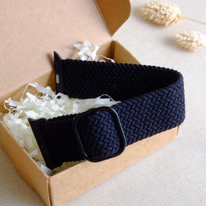 May include: A black braided watch strap with a black metal buckle. The strap is in a cardboard box with white tissue paper.