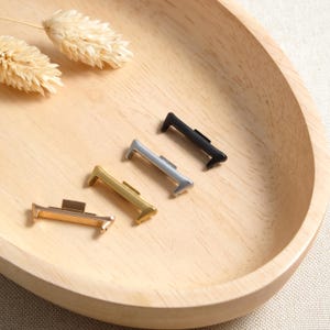 Pode incluir: Quatro adaptadores de pulseira de relógio em metal com diferentes acabamentos, incluindo ouro rosa, ouro, prata e preto. Cada adaptador possui uma barra de conexão retangular com uma pequena aba saliente para fixar a pulseira ao relógio.