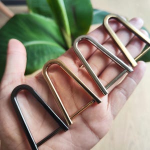 May include: A hand displays four metal U-shaped buckles in black, gold, silver, and brass. The buckles are arranged in a row, with the black buckle closest to the viewer. Green foliage forms the background.