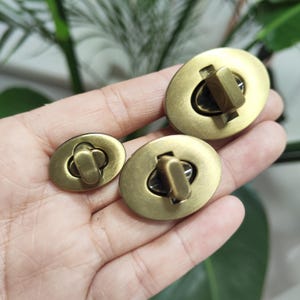 Può includere: Tre chiusure ovali per borse in ottone antico. Le chiusure hanno un design semplice con un meccanismo di bloccaggio centrale. Il metallo ha una finitura calda e invecchiata, che suggerisce un'estetica vintage o artigianale.