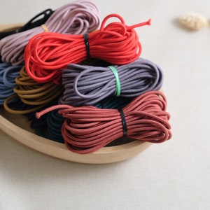 Puede incluir: Una colección de cordones elásticos de colores en un cuenco de madera. Los cordones son de varios tonos de rojo, rosa, marrón, gris y azul.