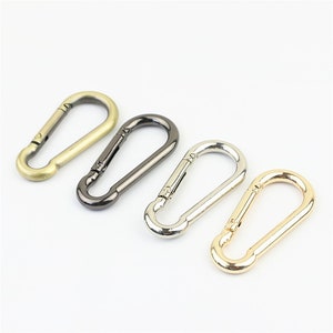 Op de afbeelding: Vier metalen sluitingen in verschillende afwerkingen: goud, zilver, zwart en antiek messing. De sluitingen zijn allemaal even groot en hebben dezelfde vorm, met een afgerond ovaal ontwerp.