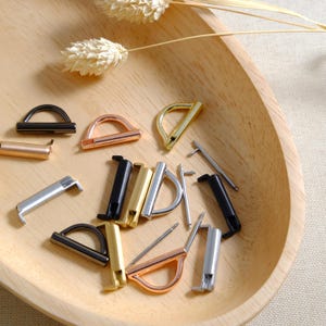 May include: Assortment of metal watch band strap connectors in various finishes, including gold, silver, black, and rose gold. The connectors are in a variety of shapes and sizes, including D-rings, bars, and spring bars.
