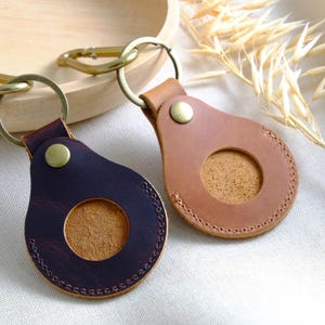Könnte beinhalten: Zwei Leder-Schlüsselanhänger in Braun und Dunkelviolett, jeweils mit einem kreisförmigen Ausschnitt und Messingbeschlägen. Die Schlüsselanhänger haben einen genähten Rand und eine Messingniete. Ein Schlüsselanhänger hat einen goldenen Karabinerhaken.