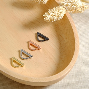 May include: Four metal D-ring clasps in different finishes: gold, rose gold, silver, and black. The clasps are arranged in a row on a wooden surface.