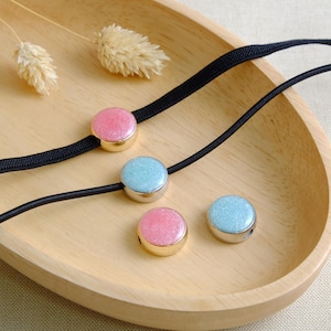 May include: Four round, sparkly, decorative beads in pink and blue. The beads are strung on a black elastic cord. The beads are on a wooden surface.