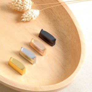 Puede incluir: Cuatro pequeñas piezas de metal decorativas en diferentes colores: negro, dorado, plateado y oro rosa. Las piezas están dispuestas en fila sobre una superficie de madera.