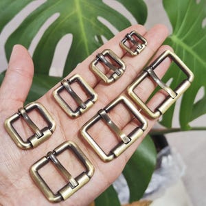 Puede incluir: Una colección de hebillas de rodillo de color latón antiguo en varios tamaños. Las hebillas son cuadradas con una barra central y un rodillo. El metal tiene un aspecto ligeramente envejecido, lo que sugiere un estilo vintage o rústico. Las hebillas se muestran en una mano.