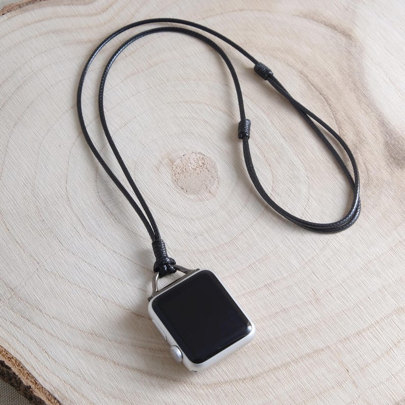 May include: A silver smartwatch with a black screen hangs from a black cord necklace. The necklace is knotted at the top and bottom, and the watch rests on a light brown wooden surface.