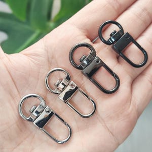 Può includere: Un set di quattro chiusure girevoli in metallo argentato e nero. Le chiusure hanno un anello girevole circolare e un gancio rettangolare. Adatte per portachiavi, borse o altri accessori.