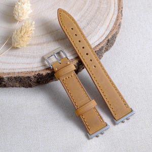 Könnte beinhalten: Ein hellbraunes Lederarmband mit silberner Schnalle und Beschlägen. Das Armband besteht aus glattem, hellbraunem Leder und hat eine genähte Kante. Das Armband ist etwa 23 cm lang.