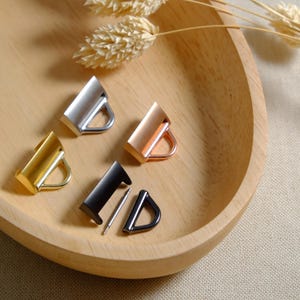 Puede incluir: Cuatro hebillas deslizantes de metal en diferentes acabados: plata, oro, oro rosa y negro. Las hebillas se muestran sobre una superficie de madera.